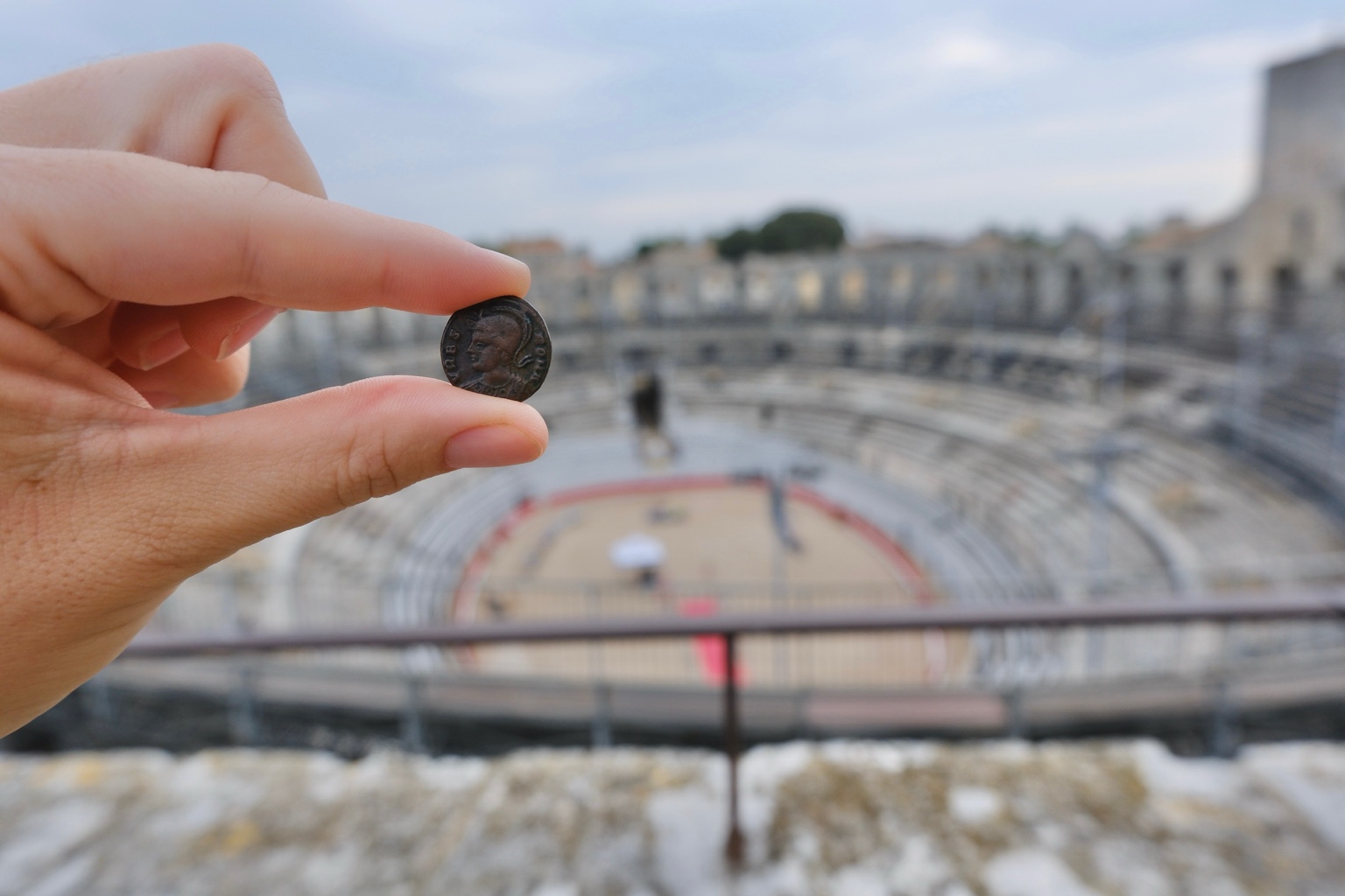The collection’s VRBS ROMA coin held between two fingers, with the arena and seating of the Arles amphitheatre beyond.