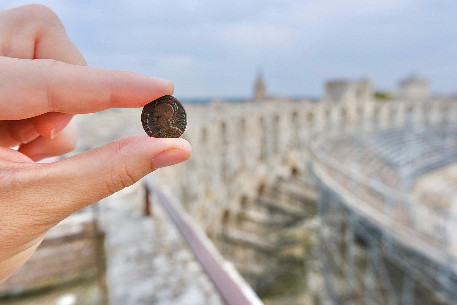 The Roma side of the coin held before the walls and seating of the Arles amphitheatre.
